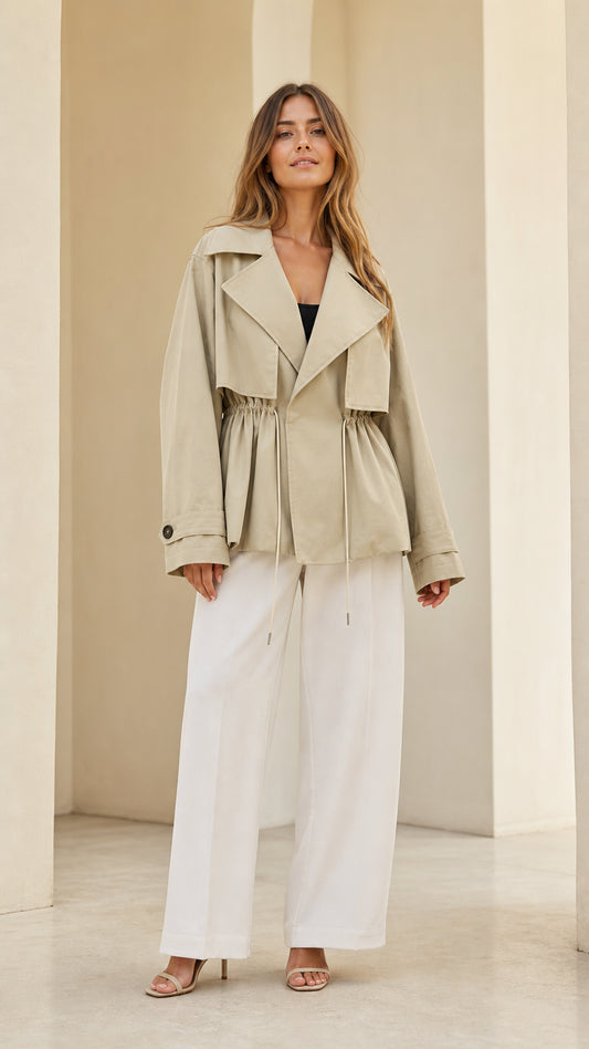 Full front view of model wearing light khaki short trench jacket with cinched drawstring waist and white wide-leg trousers